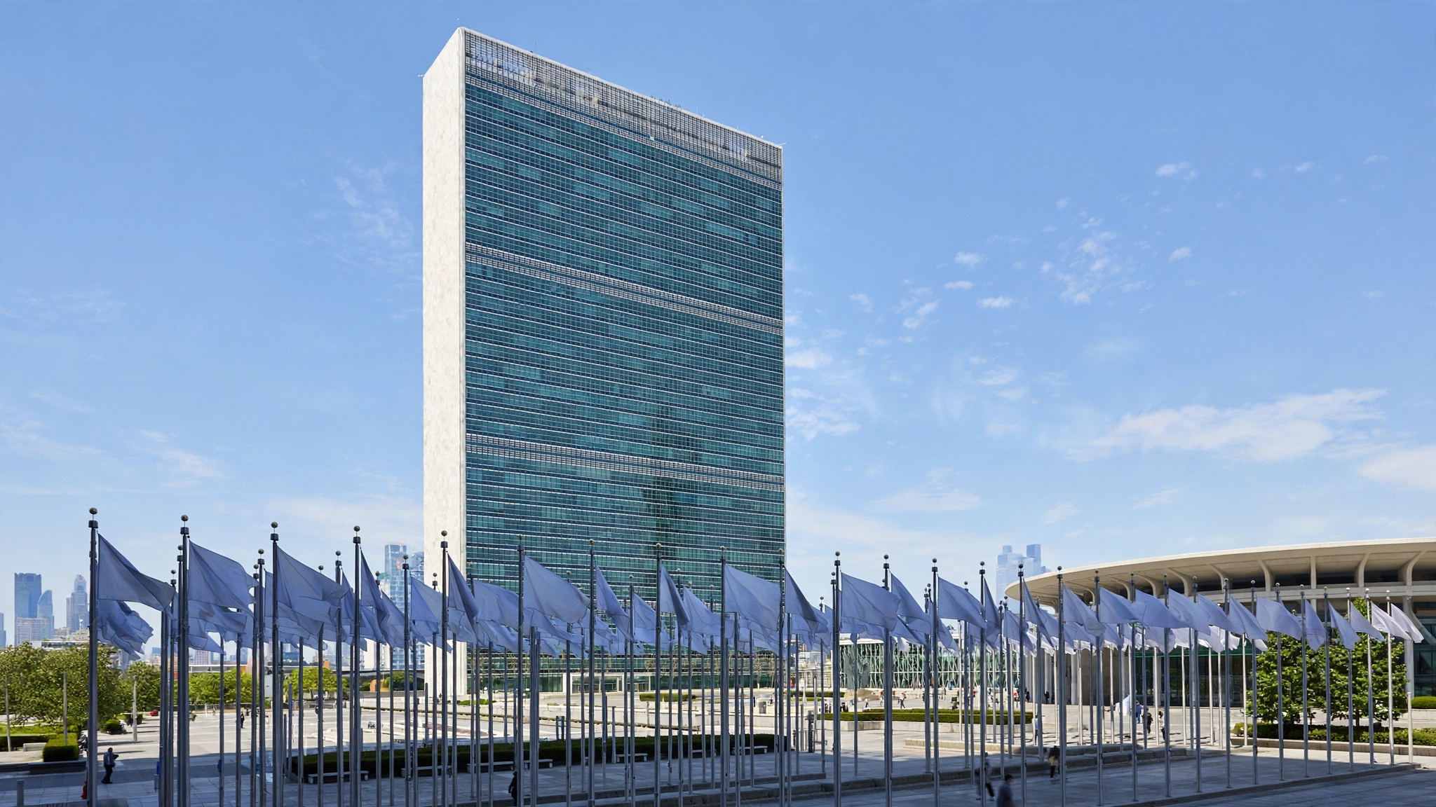 The United Nations headquarters building in New York with rows of member-state flags flying in front of it.